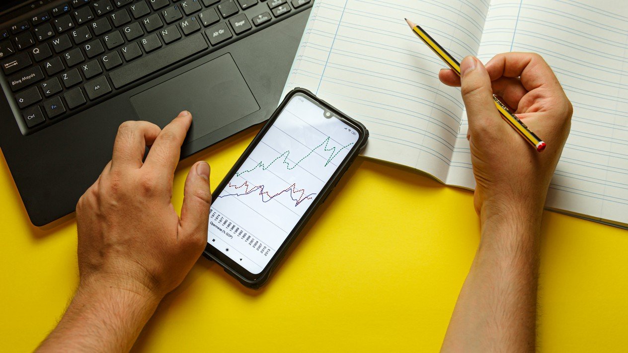 Hands using a laptop trackpad and holding a pencil over an open lined notebook, beside a smartphone displaying line graphs on a yellow desk