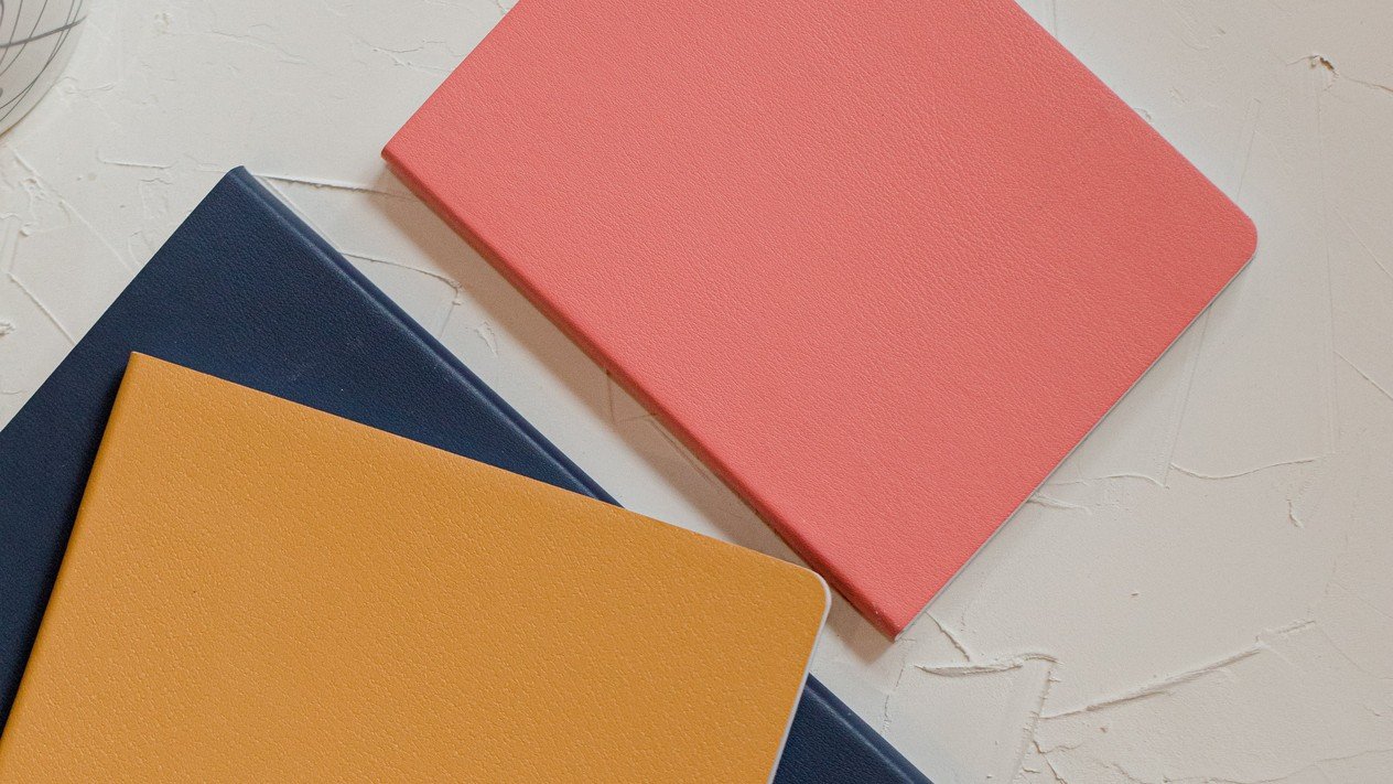 Coral-pink, mustard-yellow and navy notebooks on a textured off-white surface